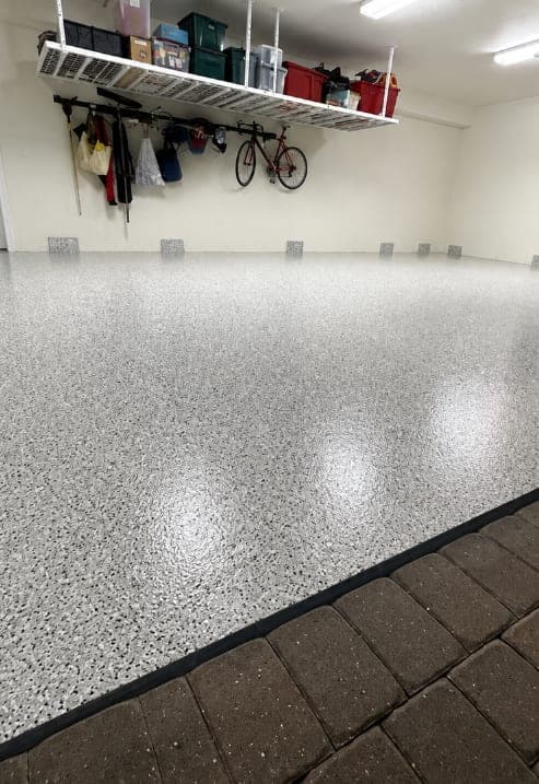 Light grey and white flake garage floor beneath an overhead storage rack holding bins and a bicycle, with brick pavers visible through the open door.