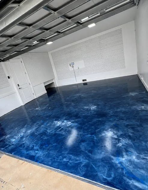 Deep blue marbled metallic epoxy garage floor beneath white walls, a ceiling storage rack, and a wall-mounted fan.